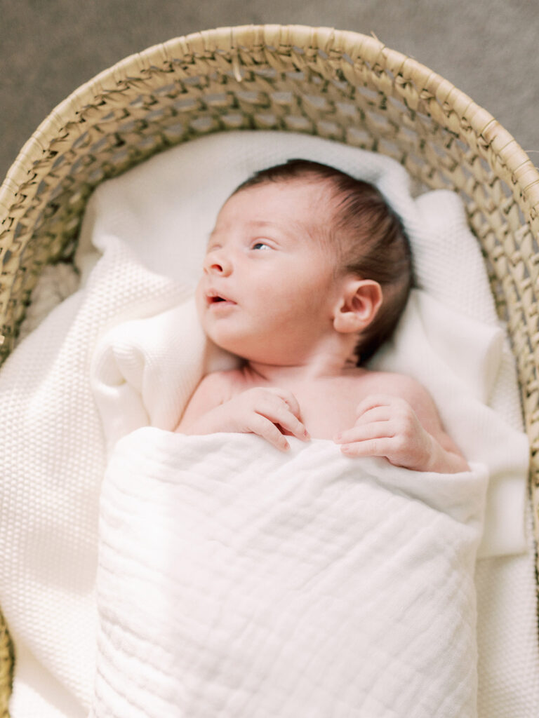A baby looking to his right while wrapped in a blanket and laying in a basket during in-home newborn photos by Katie Stansfield Photography.