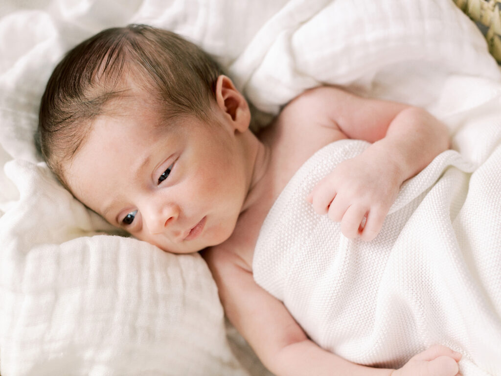 A baby wrapped in a blanket and looking to his right while laying on a blanket during in-home newborn photos by Katie Stansfield Photography.