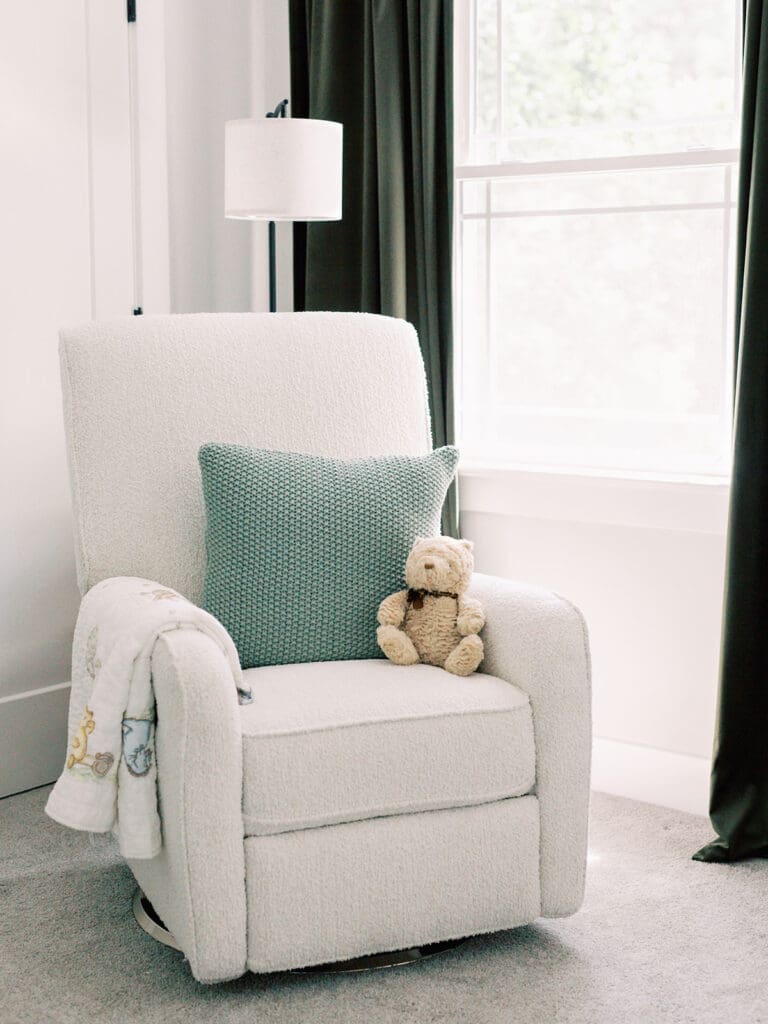 A white glider with a white Winnie the Pooh quilt, green crocheted pillow, and stuffed bear next to a window with dark green curtains at a Richmond newborn nursery photography session by Katie Stansfield Photography.
