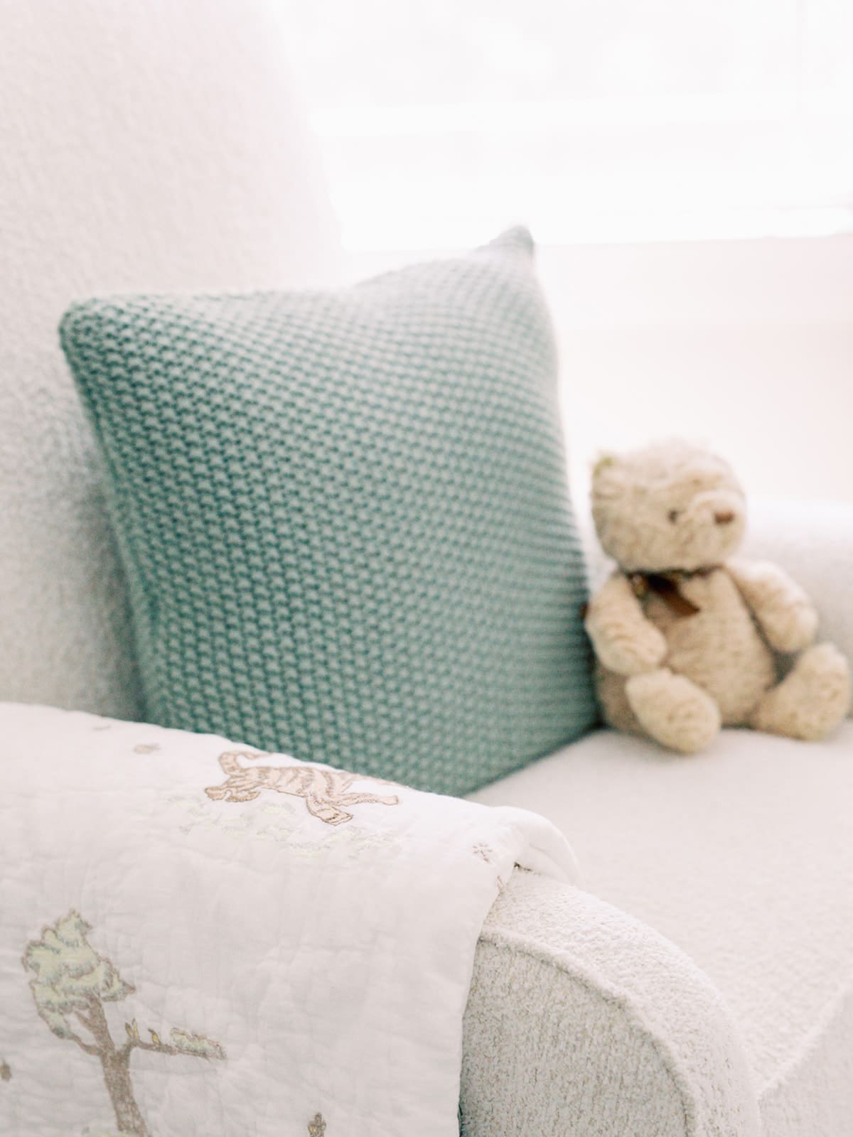 A green crocheted pillow, white quilt, and stuffed bear on a white glider at a Richmond newborn nursery photography session by Katie Stansfield Photography.