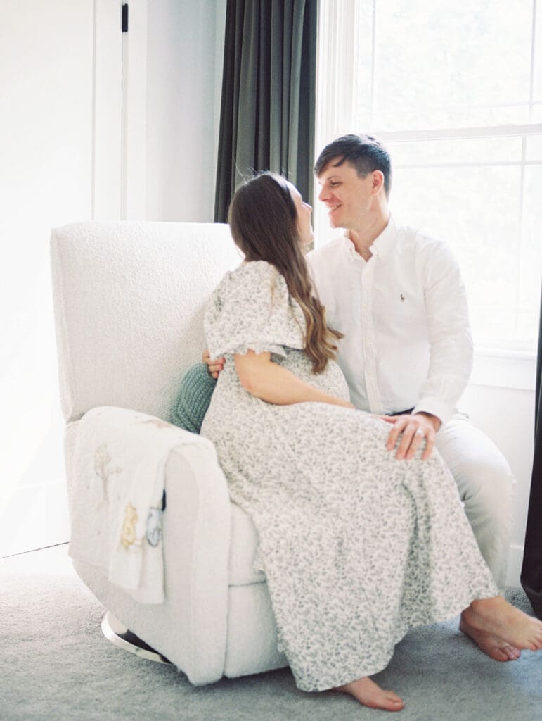 A couple smiling at each other by a window with the woman sitting in a chair and the man kneeling beside the chair at a Richmond newborn nursery photography session by Katie Stansfield Photography.