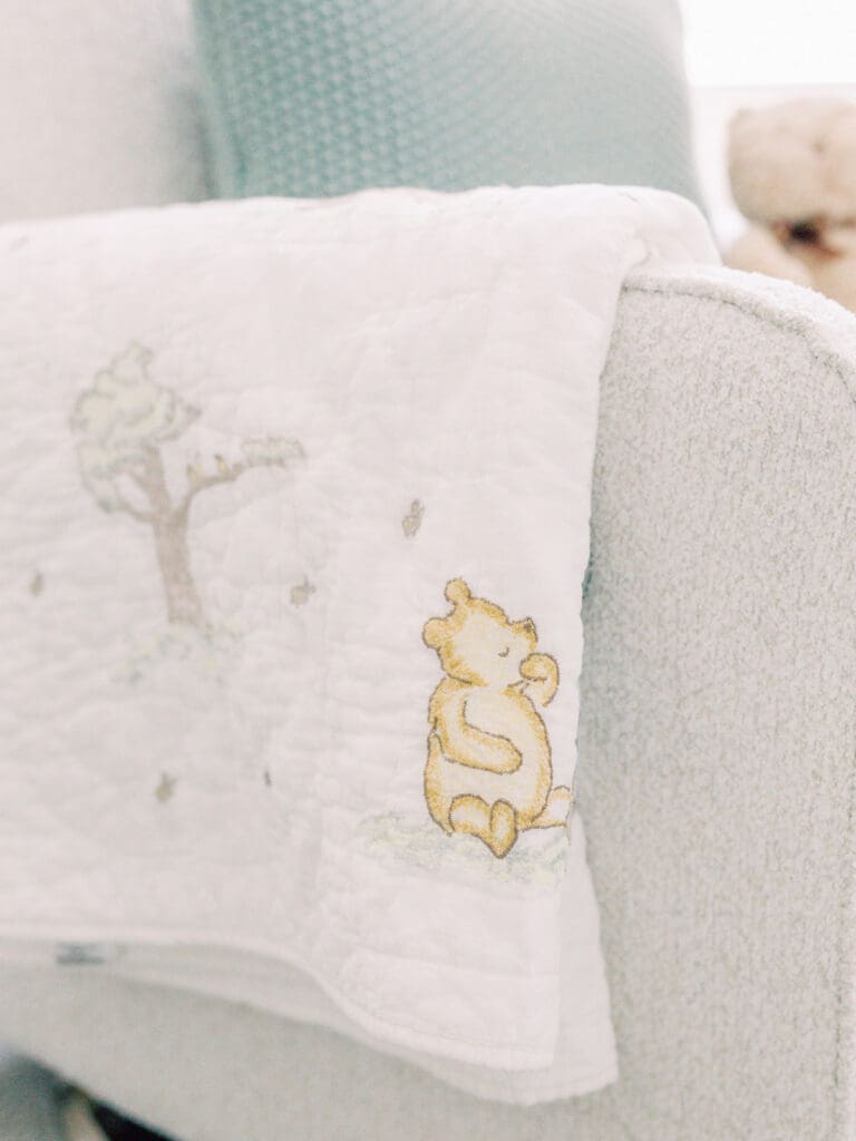 A close-up of a white Winnie the Pooh quilt on a white glider with a green crocheted pillow and stuffed bear in the background at a Richmond newborn nursery photography session by Katie Stansfield Photography.