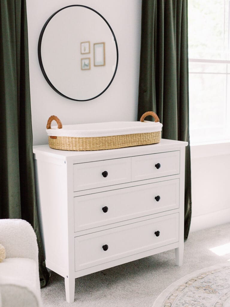 A white dresser, woven changing basket, round mirror, and dark green curtains at a Richmond newborn nursery photography session by Katie Stansfield Photography.