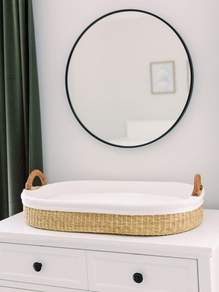 A woven changing basket on a white dresser with a round mirror on the wall at a Richmond newborn nursery photography session by Katie Stansfield Photography.