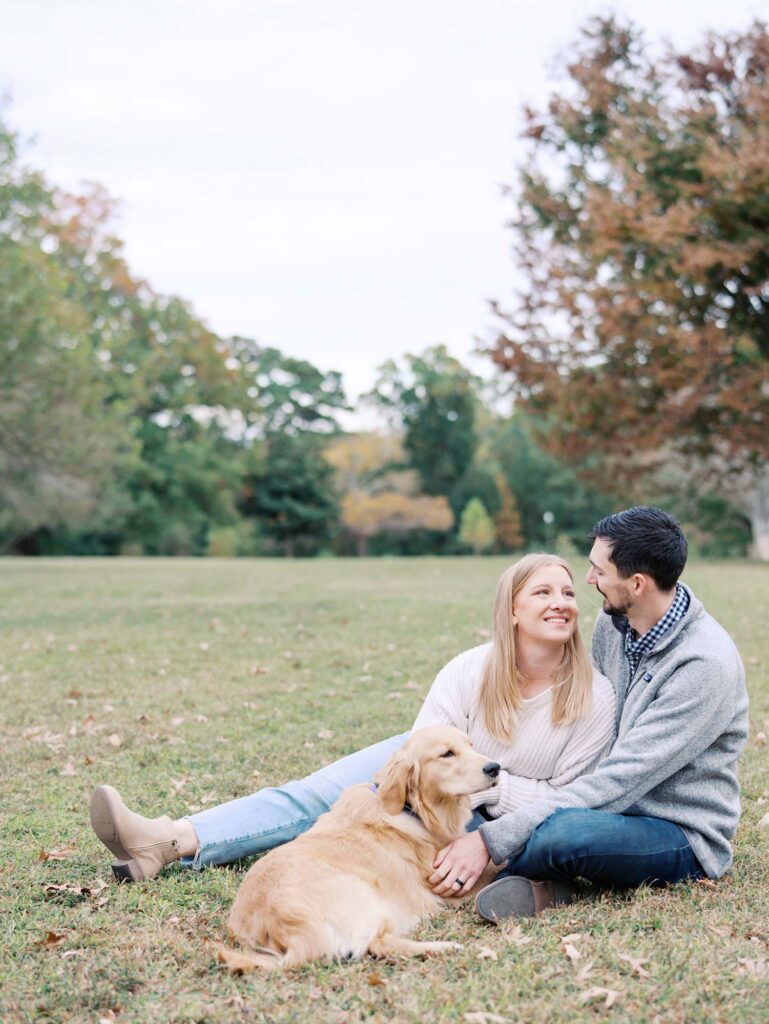 A couple smiling at each other with their dog at Forest Hill Park, one of the best locations for Richmond fall family photos, by Katie Stansfield Photography.