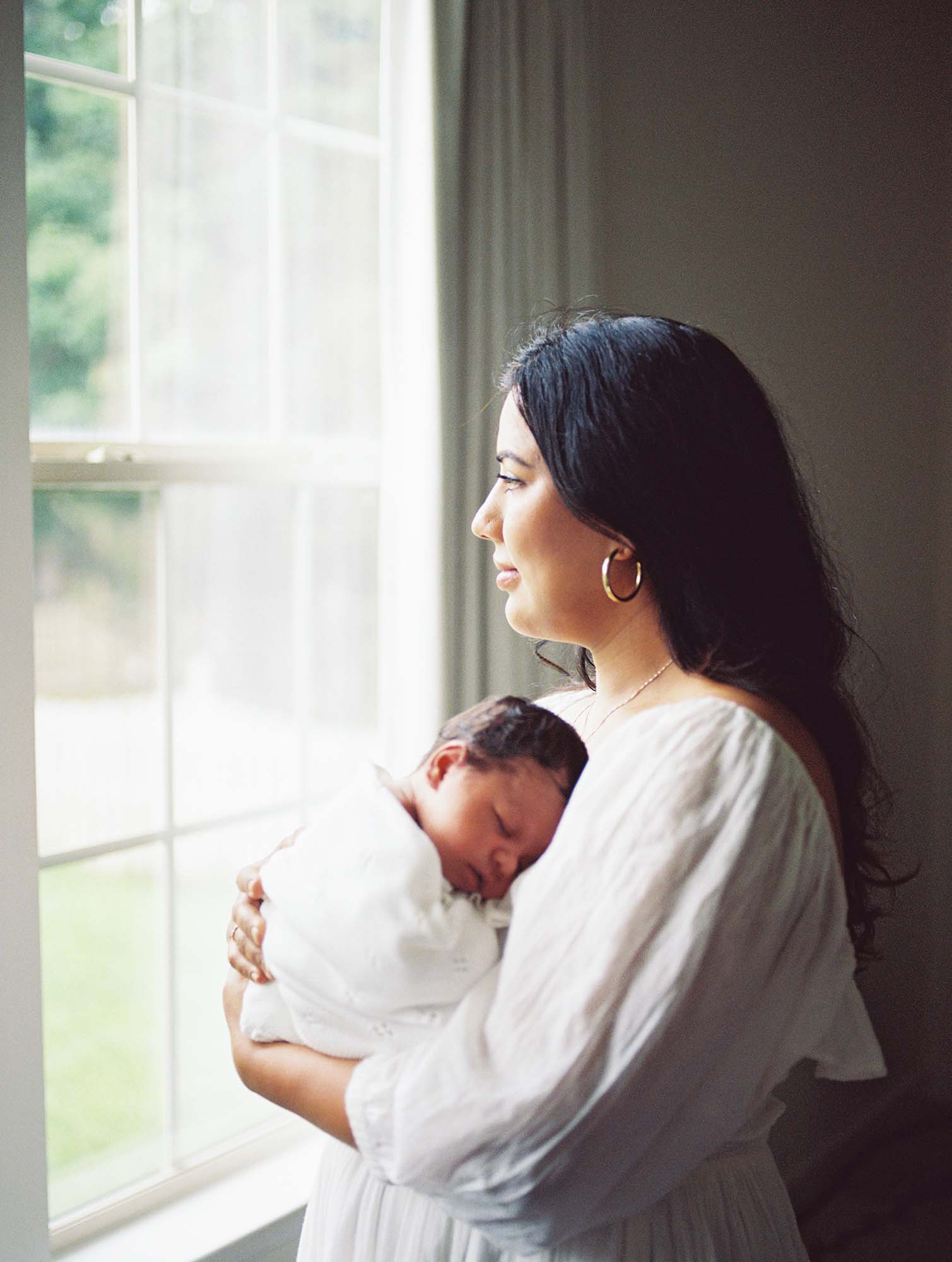 A mom holding her baby while looking outside of a window by Katie Stansfield Photography, a newborn photographer in Virginia.