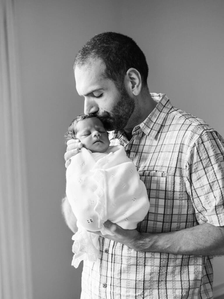 A dad holding a baby wrapped in a blanket and kissing the baby's head during a newborn photography session in Richmond, Virginia by Katie Stansfield Photography.