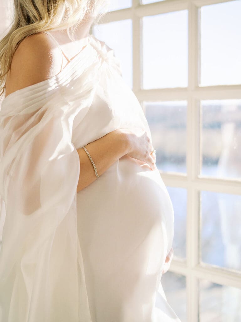 A close-up of an expecting mom holding her baby bump by a window at a maternity photography session by Katie Stansfield Photography.
