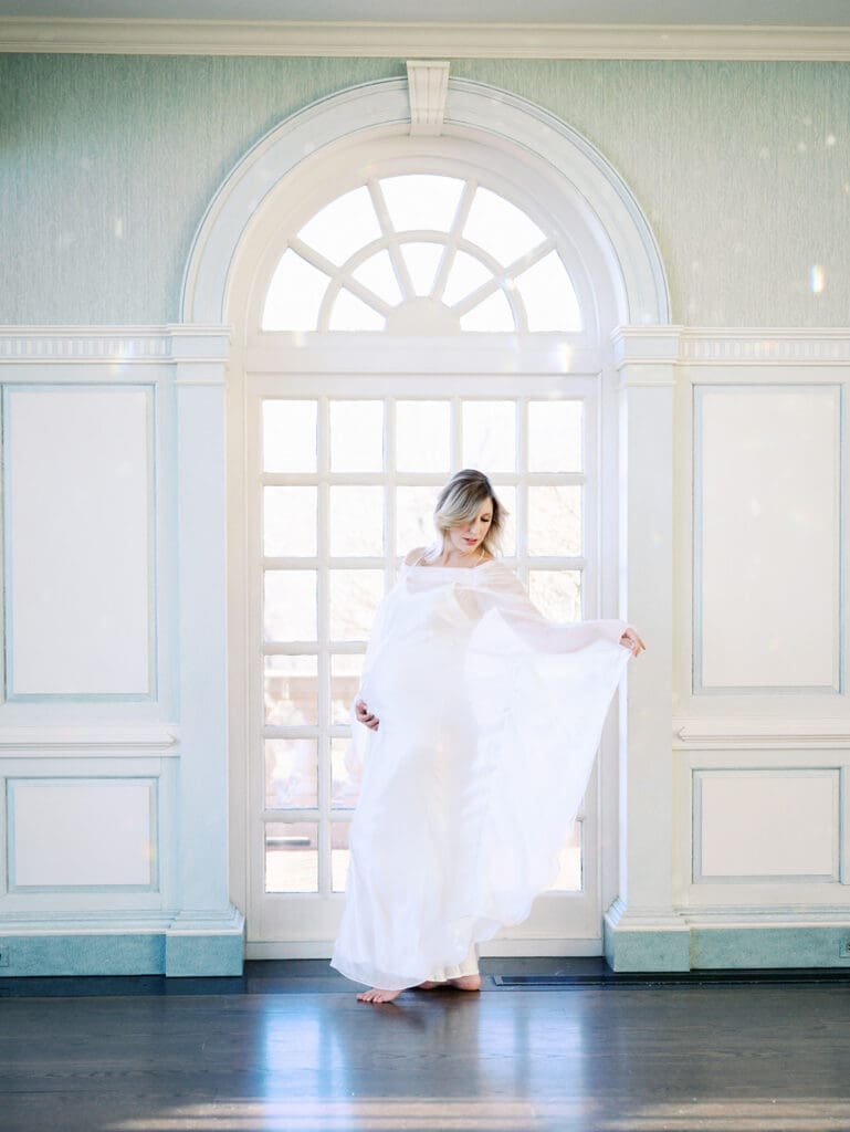 An expecting mom holding her baby bump while posing by a window at an indoor photoshoot location in Richmond by Katie Stansfield Photography.
