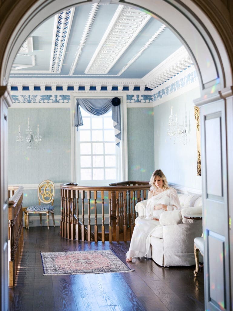 An expecting mom holding her baby bump while sitting on a sofa in a decorative room at an indoor photoshoot location in Richmond by Katie Stansfield Photography.