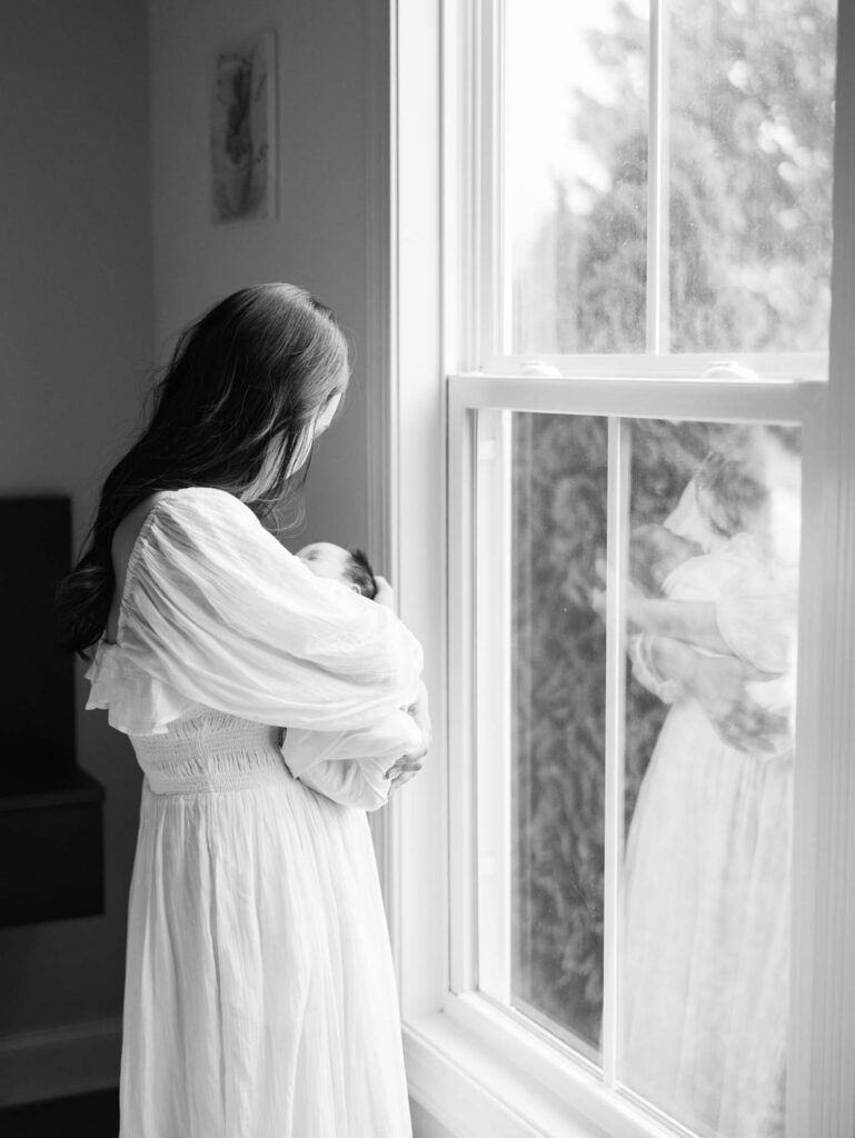 A looking down at her baby while standing by a window at an in-home session by Katie Stansfield Photography, a Midlothian newborn photographer.