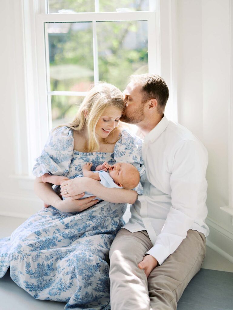 A dad kissing mom's head as the mom looks down at the baby at a newborn session by Katie Stansfield Photography.