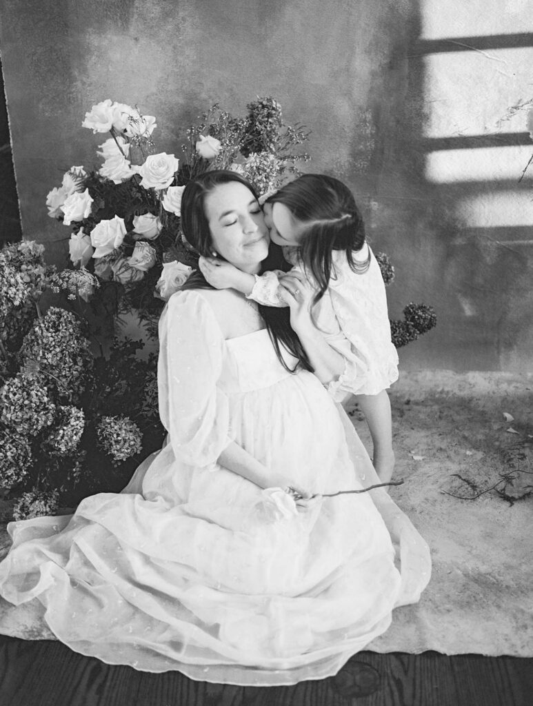 A young girl hugging and kissing her mom at an indoor maternity photography session by Katie Stansfield Photography.
