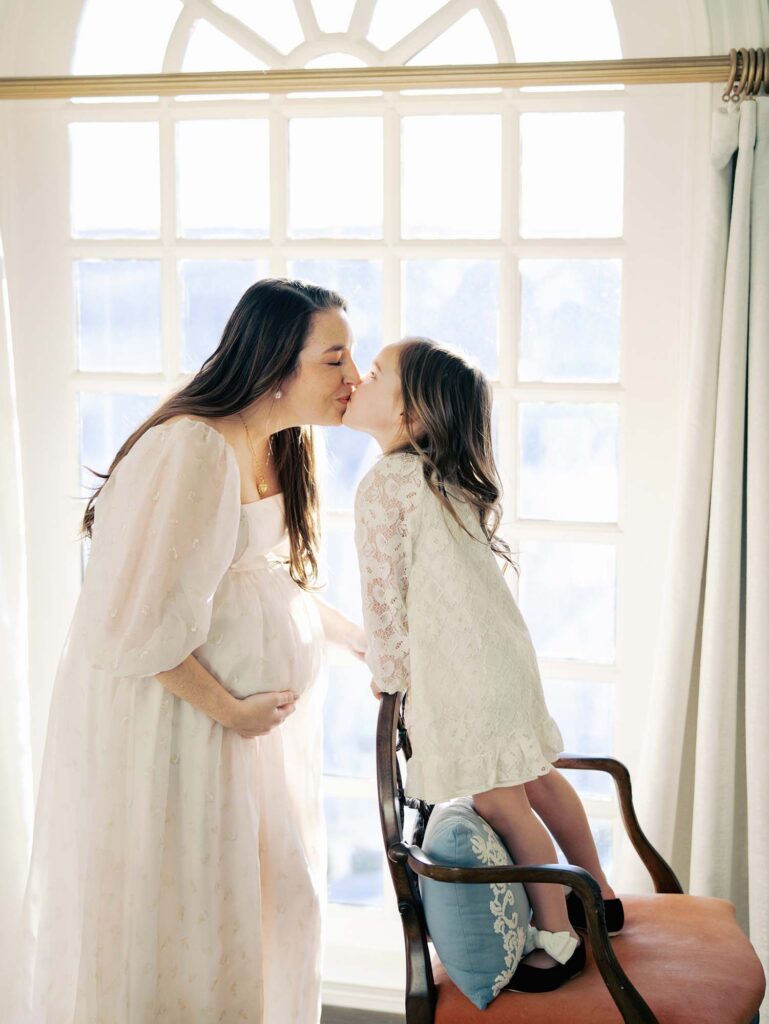 A mom and her daughter kissing by a window at a maternity photography session by Katie Stansfield Photography.