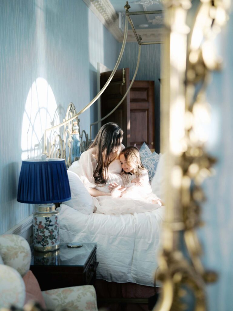 A mirror reflection of a mom and daughter sitting on a bed at a maternity session by Katie Stansfield Photography, a Bon Air maternity photographer.
