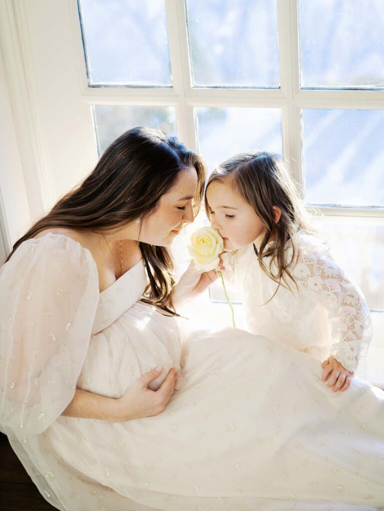 A mom and her daughter smelling a flower as the mom holds her baby bump at a bon air maternity photography session by Katie Stansfield Photography.