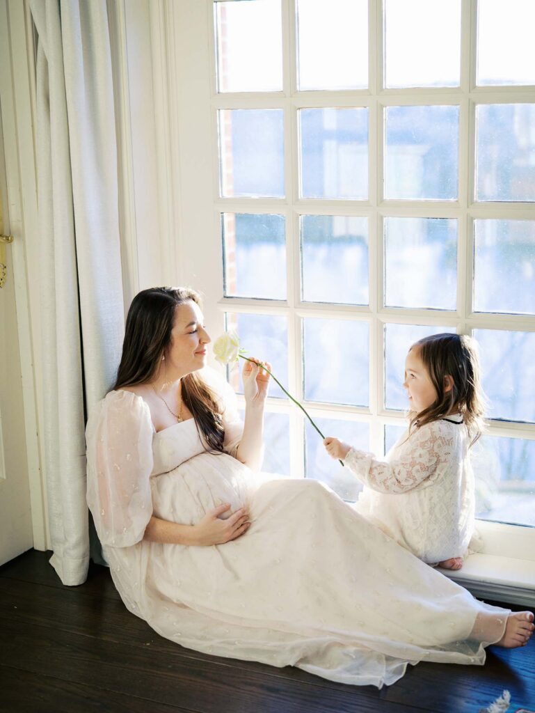 A daughter holding out a flower for her mom to smell as the mom holds her baby bump at a maternity session by Katie Stansfield Photography, a Bon Air maternity photographer.