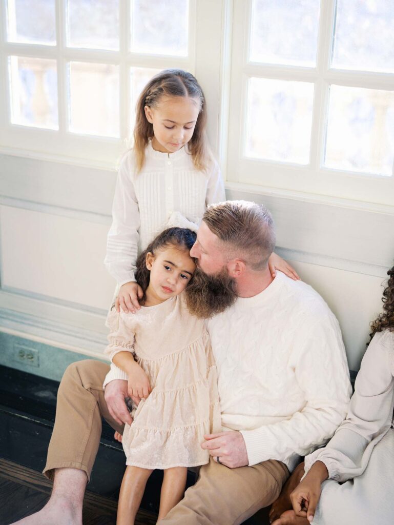 A dad kissing his daughter as another daughter smiles at them to show how to choose a photographer for your family in Richmond, VA by Katie Stansfield Photography.
