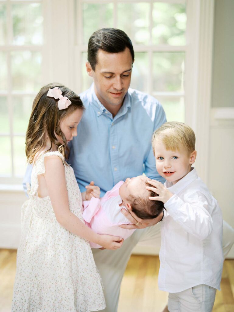 A dad kneeling while holding a baby as sister looks at the baby and brother smiles in their home, one of the newborn photo locations in Richmond VA, by Katie Stansfield Photography.