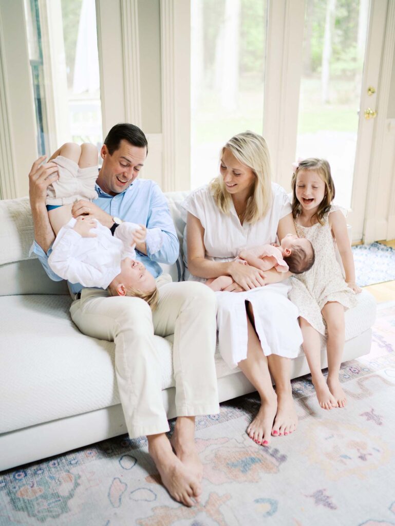 A family sitting on a sofa in their home, one of the newborn photo locations in Richmond VA, by Katie Stansfield Photography. The dad is tickling brother, mom is holding the baby, and sister is laughing.