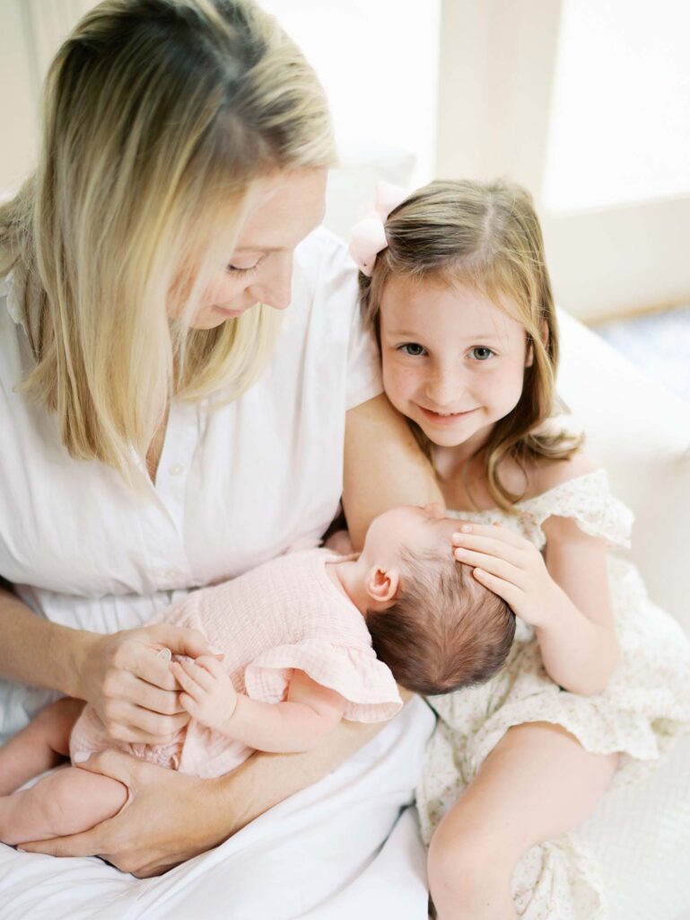 A mom holding a baby as the sister smiles and touches baby's head at a newborn session by Katie Stansfield Photography.