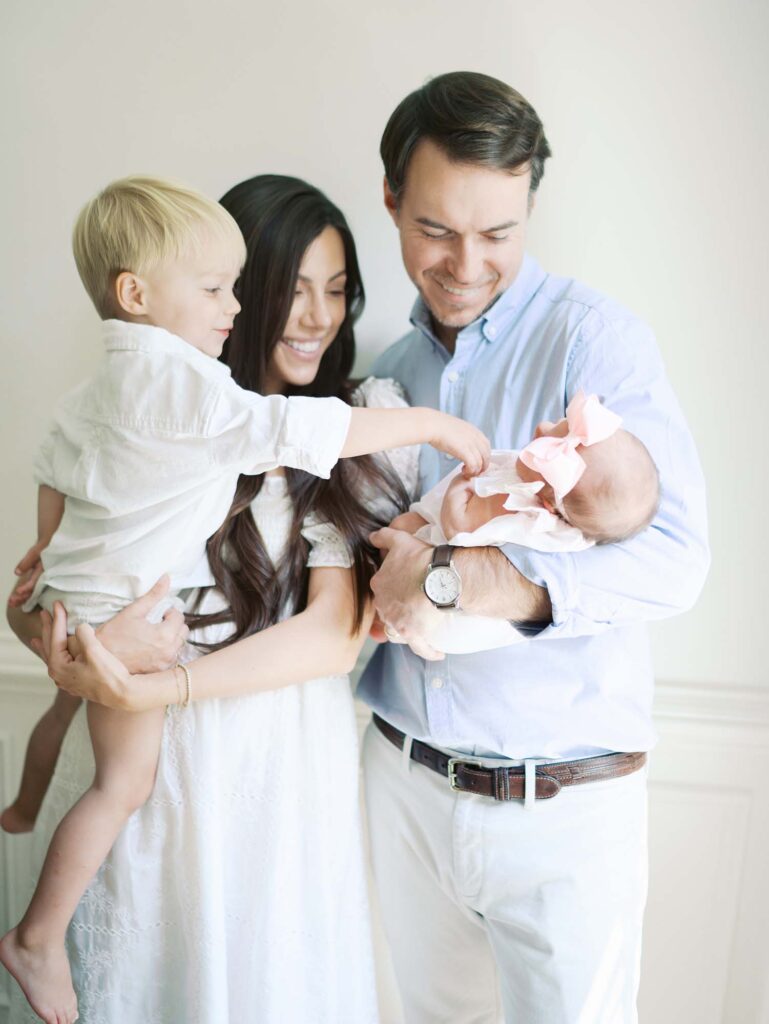 A dad holding a baby and mom holding a toddler as the toddler points at the baby at a newborn session by Katie Stansfield Photography.