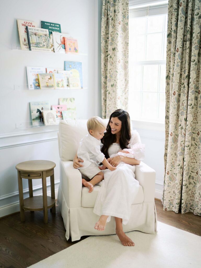 A mom in a nursery holding her baby and smiling at her toddler as the toddler looks at the baby by Katie Stansfield Photography, a Bon Air newborn photographer.