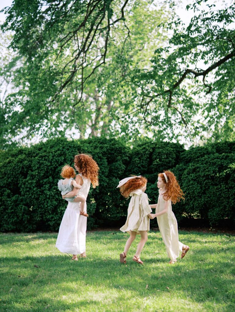 A mom holding a toddler as her two older daughters hold hands and jump in a circle at a family session by Katie Stansfield Photography.