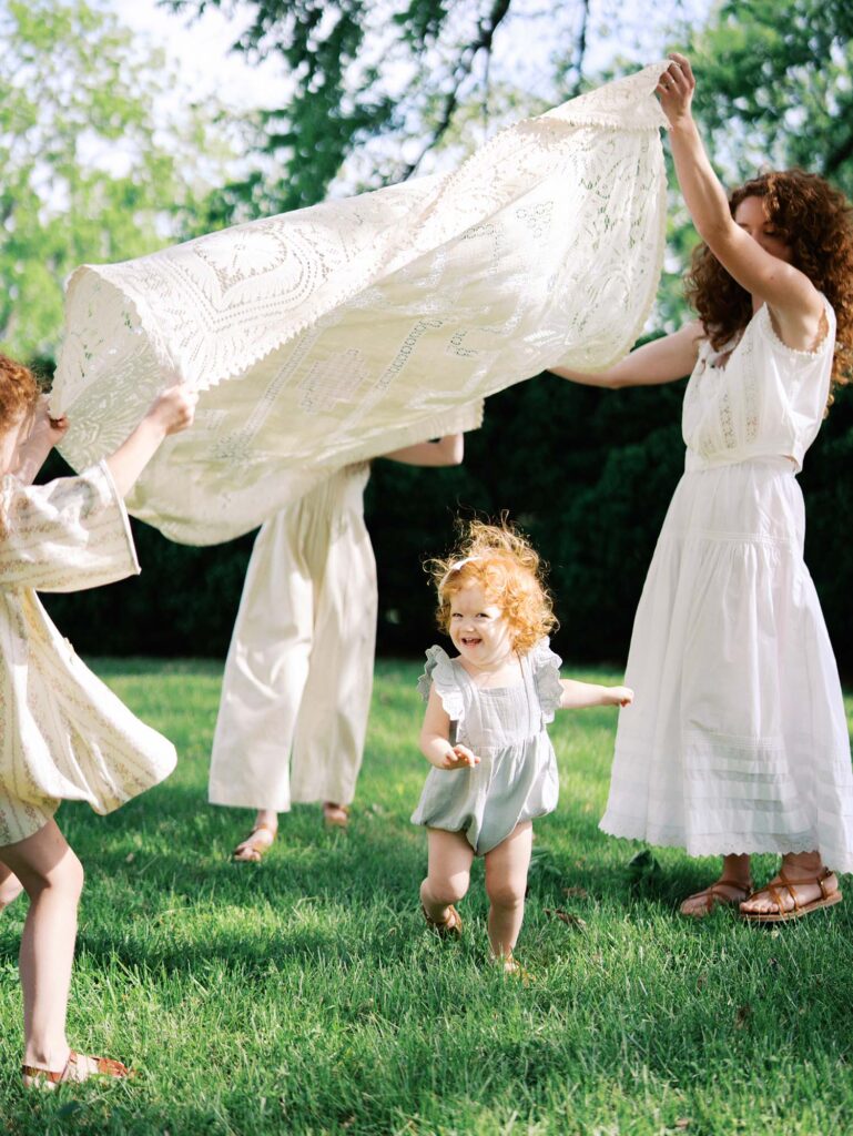 A family holding up a blanket as a toddler runs underneath by Katie Stansfield Photography, a family photographer in Short Pump VA.