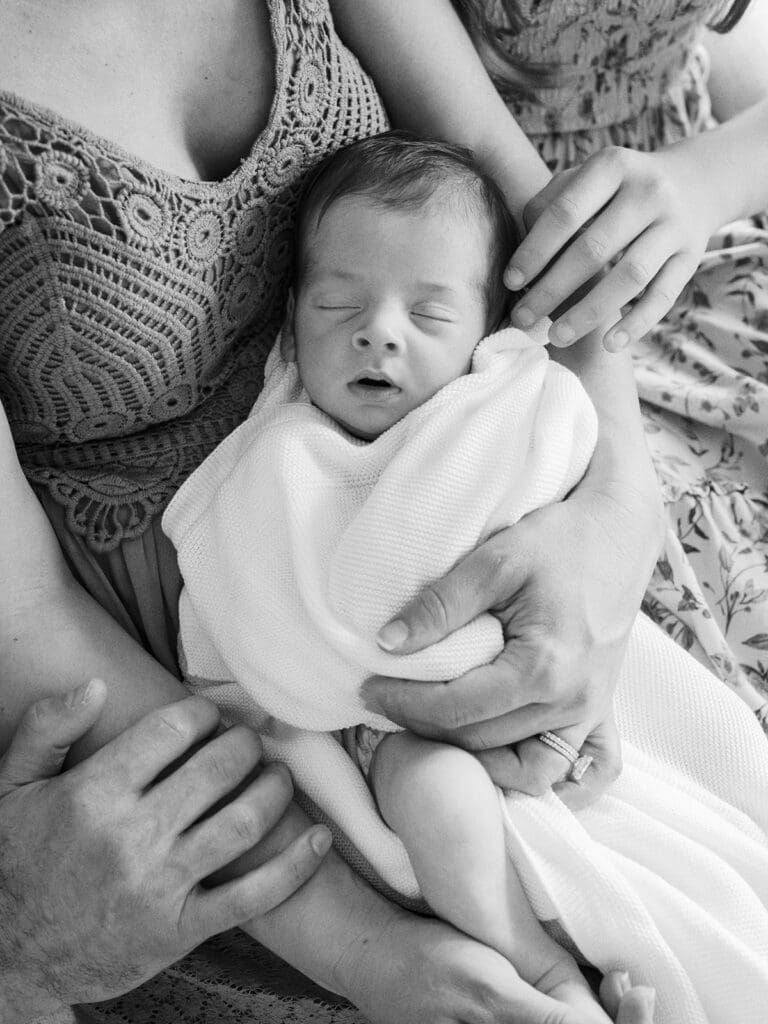 A baby wrapped in a blanket while being held by the mother and other family members' hands touching the mother's arms at a Richmond in-home newborn photography session by Katie Stansfield Photography.