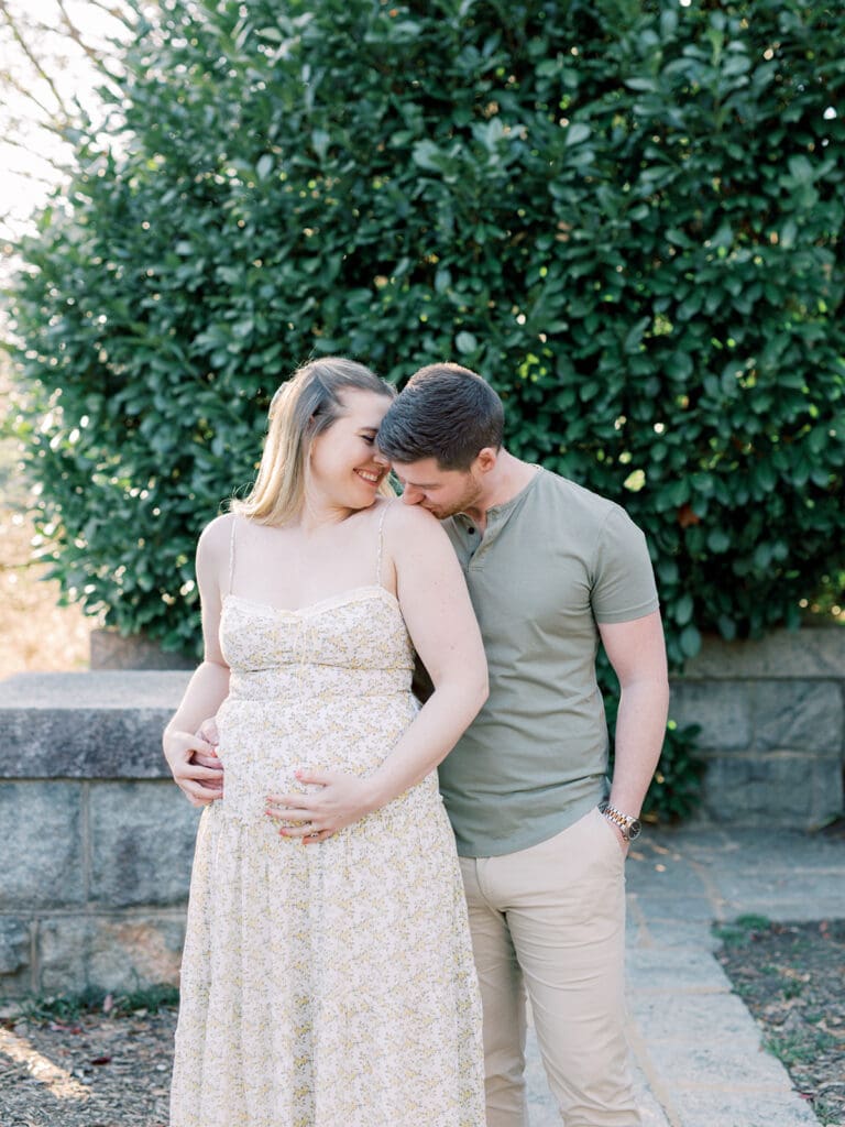 A woman smiling while looking at a man and holding her bump as the man kisses her shoulder during a maternity photography in Virginia session by Katie Stansfield Photography.