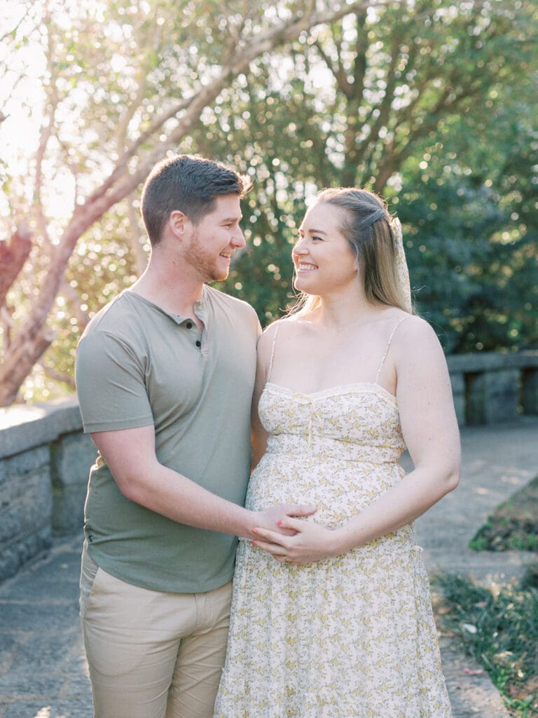 A man and woman smiling at each other while holding the woman's pregnancy bump during a Richmond maternity photography session by Katie Stansfield Photography.