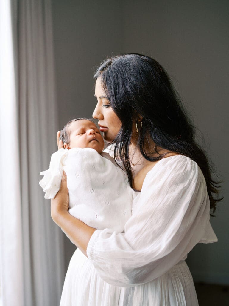 A mom in a white dress leaning in to kiss a baby's temple as the baby is wrapped in a white blanket during a newborn photography session in Richmond, Virginia by Katie Stansfield Photography.