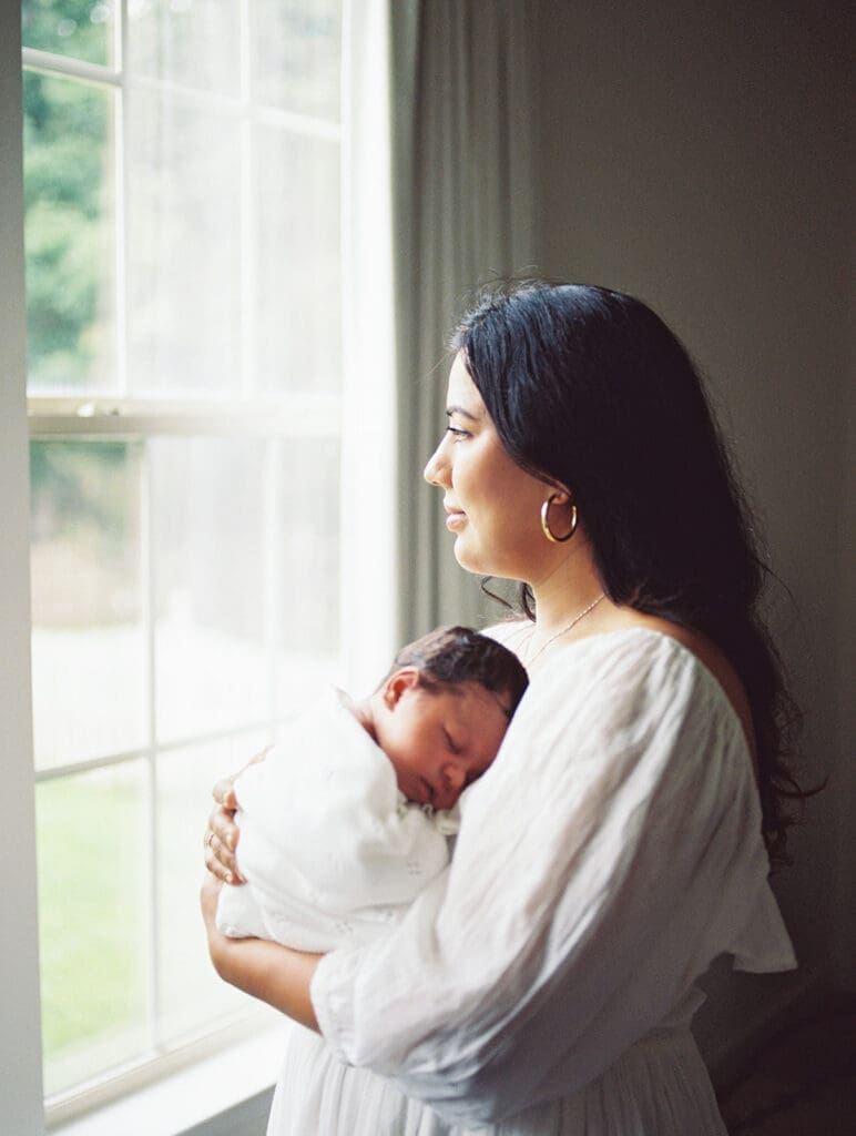 A mom holding a baby and looking out of the window during a newborn photography session in Richmond, Virginia by Katie Stansfield Photography.