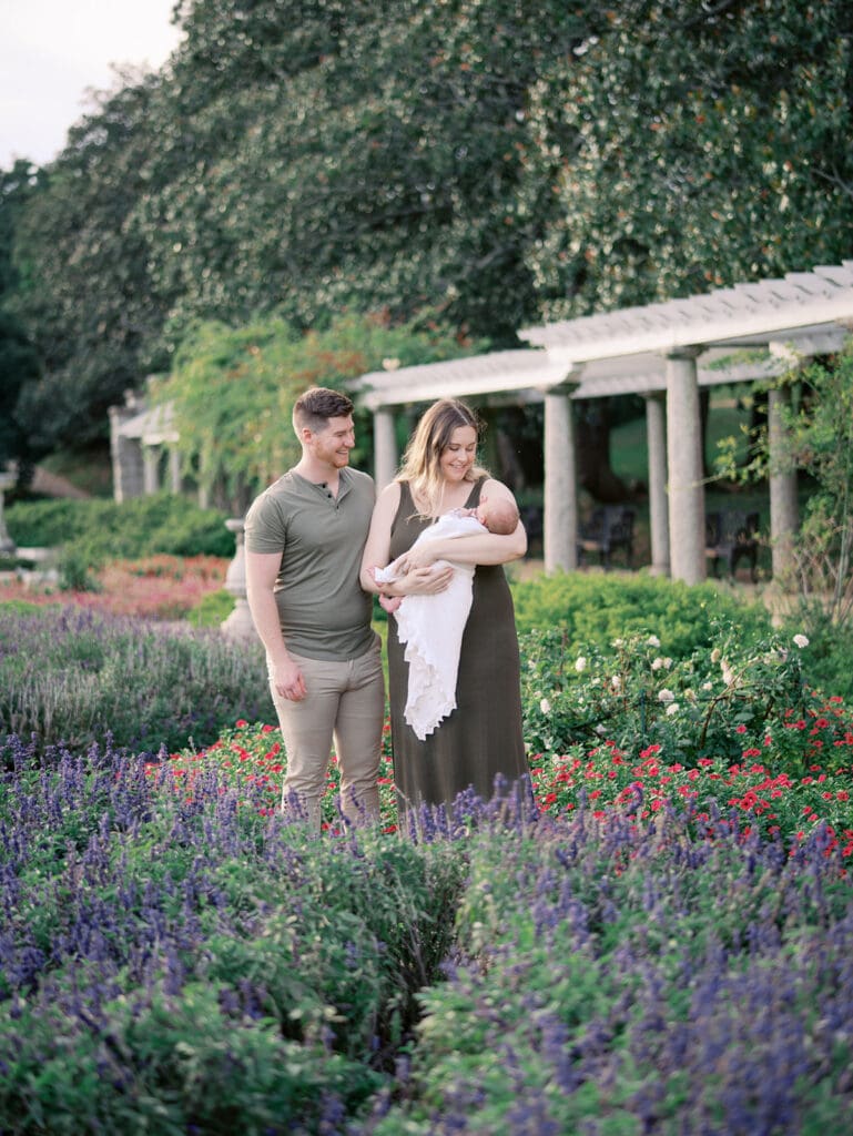 A mom and dad standing in a garden as mom holds their baby during a newborn session by Katie Stansfield Photography.
